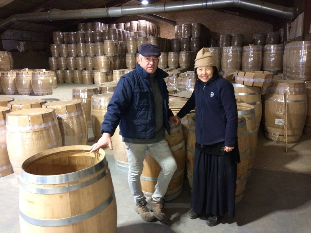 Two people standing next to barrels in a wine cellar.